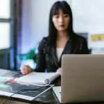 Giovane ragazza segue una lezione a distanza seduta alla scrivania con davanti a lei un computer portatile e uno smartphone usato come webcam
