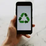 Mano in primo piano sorregge uno smartphone che mostra il simbolo universale del riciclo con tre frecce verdi che formano un triangolo